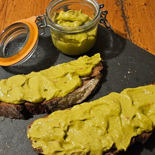 Deux tranches de pain grillé tartiné avec la tartinade d'avocat à la tomate. Elles sont posées à côté du pot de tartinade, sur un plat en ardoise.